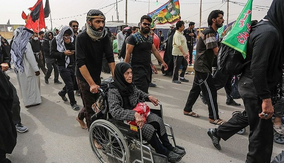 تصویر یک پیرزن مسلمان نشسته روی ویلچر در مسیر حرم امام حسین در مراسم مذهبی پیاده روی اربعین که یک مرد جوان در حال هل دادن ویلچر است. در اطراف تعداد زیادی مرد و زن در حال پیاده روی هستند
